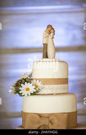 Dreistöckige Erdhochzeitstorte mit Leinwandspitze und Schleifen und Sonnenblumen auf Holzrindenständer und Holztürhintergrund Stockfoto