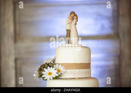 Dreistöckige Erdhochzeitstorte mit Leinwandspitze und Schleifen und Sonnenblumen auf Holzrindenständer und Holztürhintergrund Stockfoto