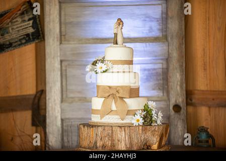 Dreistöckige Erdhochzeitstorte mit Leinwandspitze und Schleifen und Sonnenblumen auf Holzrindenständer und Holztürhintergrund Stockfoto