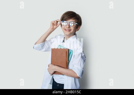 Porträt eines kaukasischen Jungen in weißem Hemd mit einem Stapel von Kopierbüchern, der lächelnd steht und seine Brille auf weißem Hintergrund hält Stockfoto