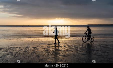 Silhouette Bild von zwei Menschen laufen und Radfahren auf einem Sandstrand bei Sonnenaufgang Stockfoto