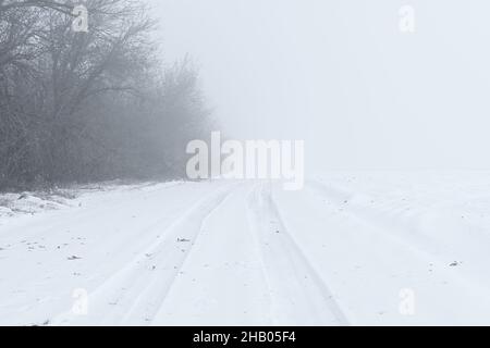 Winterlicher Schneefall auf der Straße. Eine leere Landstraße ohne Autos, bedeckt mit Schneeverwehungen. Reisen Sie bei schlechtem Wetter. Winter wolkige Landschaft mit einer Landstraße. T Stockfoto