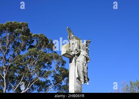 Steinskulptur eines Engels vor einem Kreuz, wobei das himmlische Wesen auf den strahlend blauen Himmel zeigt Stockfoto