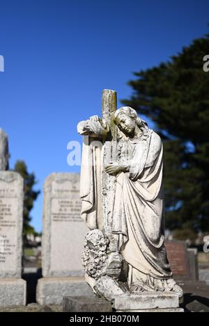 Porträt einer Steinskulptur einer Frau, die auf einem Friedhof über ein christliches Kreuz drapiert ist, mit Gräbern im Hintergrund Stockfoto