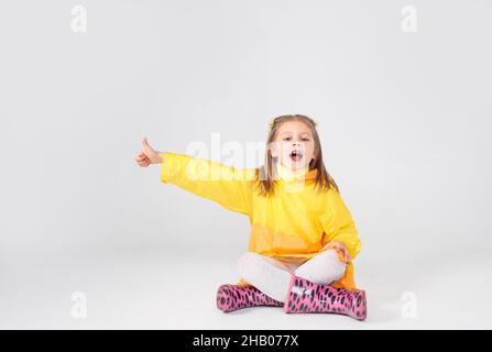 Ein kleines Mädchen in einem gelben Regenmantel und Stiefeln sitzt auf einem weißen Hintergrund und zeigt ihren Finger nach oben. Stockfoto