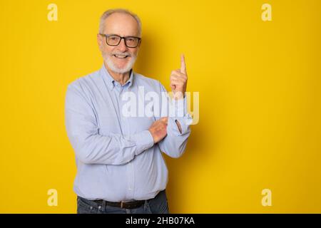 Älterer grauhaariger Mann mit elegantem Hemd, der auf einem isolierten gelben Hintergrund steht und mit dem Finger Nummer eins nach oben zeigt und dabei mit einem Lächeln auf den Lippen lächelt Stockfoto