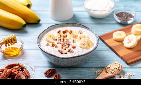 Über Nacht Hafer mit Banane und Pekannüsse, Chiasamen in Schüssel mit Zutaten herum. Gesundes proteinreiches Frühstück auf blauem Holztisch, Draufsicht Stockfoto