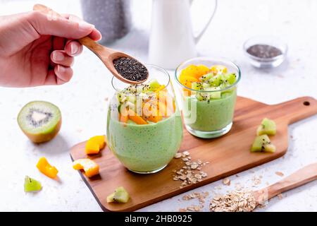 Frau, die Chiasamen zu ihrem gesunden Frühstück hinzufügt. Grüner Matcha über Nacht Hafer mit tropischen Früchten Kiwi und Mango in Gläsern Stockfoto