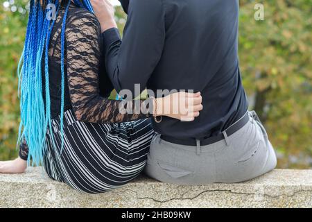 Der Mittelteil eines umarmenden heterosexuellen Paares. Blick von hinten. Junge Frau mit langen blauen Dreadlocks und jungen Mann. Die Männer sitzen auf einem Stockfoto