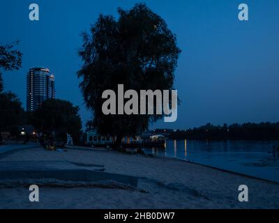 Abends Landschaft im Freien in blauen Farben mit Menschen, die sich am Strand des Flusses Dnipro in Kiew ausruhen. Schwimmendes Wasser-Restaurant, einsamer Baum. Stockfoto