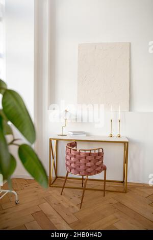 Die Inneneinrichtung des hellen Raumes ist mit einem Tisch mit Korbsessel und einer Lampe und Büchern ausgestattet, die in der Nähe einer weißen Wand mit einem weißen Wandanstrich angebracht sind Stockfoto