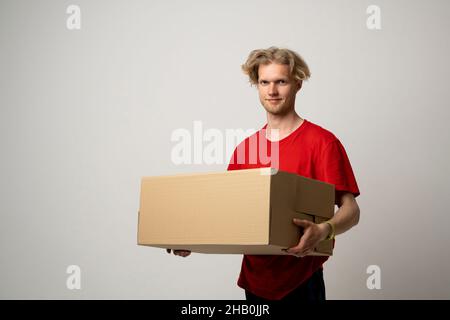 Lieferservice. Junger lächelnder Kurier mit Karton. Glücklicher junger Lieferer im roten T-Shirt, der mit Paket isoliert auf Weiß steht Stockfoto