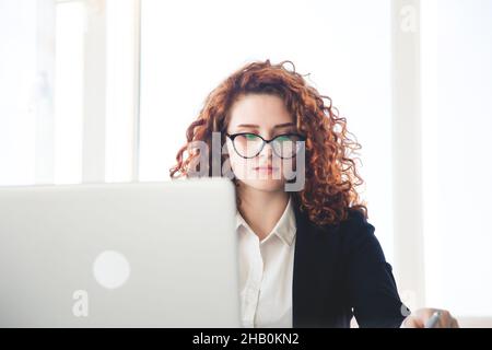 Schöne junge lockige rothaarige Frau in einem Business-Anzug und Die Brille funktioniert im Büro auf einem Laptop Stockfoto