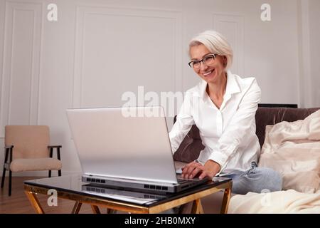 Glückliche 50 ältere ältere ältere Erwachsene Frau mittleren Alters mit Laptop mit Computer sitzen auf der Couch zu Hause. Lächelnde elegante ältere grauhaarige Dame im Glas Stockfoto