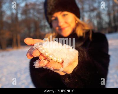 Eine junge Frau in einem schwarzen Hut und einem Nerzpelzmantel hält in ihren Handflächen eine Handvoll flauschigen weißen Schnee, in dem ein weißer Perlenschmuck liegt. Perlen und Schneeflocken funkeln in den orangen Strahlen der Kulisse Stockfoto