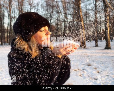 Eine junge blonde Frau in einem Pelzmantel und Hut aus Nerzpelz bläst in den orangen Strahlen der untergehenden Wintersonne leichten flauschigen Schnee von den Handflächen. Winterspaß. Stockfoto