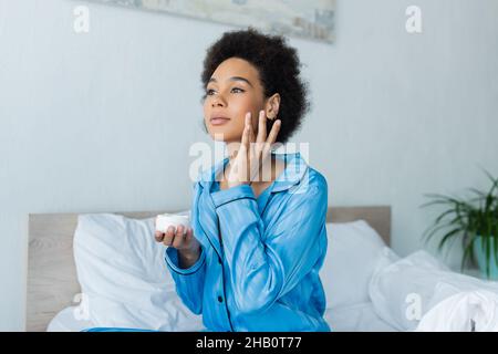 Lockige afroamerikanische Frau im Schlafanzug, der Behälter hält und Gesichtscreme im Schlafzimmer aufgibt Stockfoto