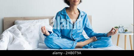 Beschnittene Ansicht einer afroamerikanischen Frau im Schlafanzug, die auf dem Bett meditiert, Banner Stockfoto