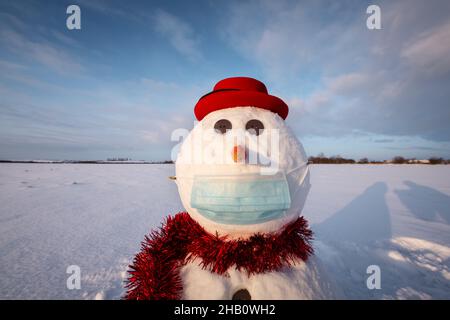 Lustiger Schneemann in stilvollem roten Hut mit medizinischer Maske auf schneebedecktem Feld. Abgesagte Reise und soziales Distanzierungskonzept Stockfoto