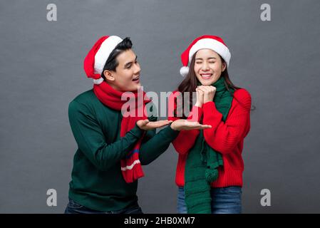 Junger, hübscher asiatischer Mann, der seine Freundin an Weihnachten überrascht, aufgenommen im Studio vor einem isolierten grauen Hintergrund Stockfoto