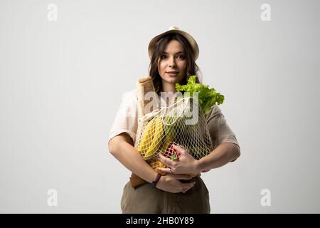 Öko Tasche Saitenbeutel mit Obst, Gemüse und Brot in den Händen eines brünetten Mädchens in einem beigen Anzug und einem Hut. Umweltfreundliches Konzept. Mode Stockfoto