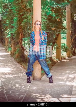 Lächelndes Mädchen mit Zöpfen in einem karierten langen Hemd übergroße Jeans und raue Stiefel stehen an der Gabel im Park, die sich auf einer Säule stützt Stockfoto