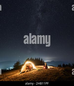 Männlicher Tourist, der Gitarre in den Händen hält widerspenstiges Lied für seine Frau, die im Zelt auf ihrem Bergcampingplatz ist. Romantische Atmosphäre des Himmels durch Sterne, Milchstraße und Licht von der Stadt unter Hügeln weit weg beleuchtet. Stockfoto