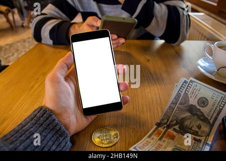 Hand hält Handy mit leerem weißen Bildschirm, während Sie in einem Café sitzen, Dollarscheine und Bitcoin auf einem Holztisch, Mockup-Bild. Stockfoto