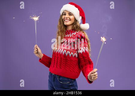 Glückliches Mädchen im Weihnachtsmann-Hut tanzt mit brennenden Wunderkerzen im Studio, das auf violettem Hintergrund gedreht wurde Stockfoto