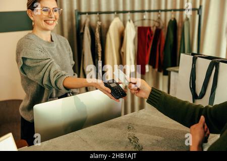 Freundliche Geschäftsassistentin, die eine Kartenzahlung von einer Kundin übernimmt. Glückliche Verkäuferin, die einer Kundin hilft, während sie hinter dem Counte steht Stockfoto