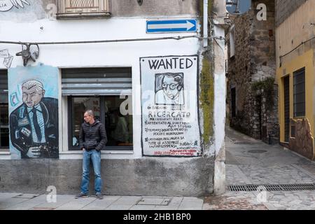 Im Dorf Orgosolo auf Sardinien steht ein Mann vor einem Fenster mit Wandmalereien an den Seiten.das Dorf Orgosolo auf Sardinien hat eine lange Tradition der politischen Straßenkunst, die an den Wänden des Dorfes sichtbar ist. Seit einigen Jahren verbinden sich die linksextremen Fresken und die Hommage an die Kämpfe des Volkes mit Gemälden zum Thema Migranten und das Coronavirus. Stockfoto