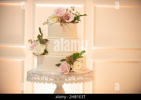Wunderschöne dreistufige Hochzeitstorte mit Perlen und rosa und weißen Blumen auf Spitzenkuchen in der Nähe der weißen Wand Stockfoto