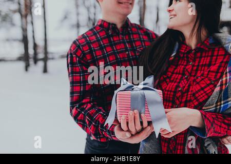 Freund gibt eine Geschenkbox Geschenk mit Band im Freien aufgrund der heiligen Valentinstag zu ihrer Freundin. Verschneite Wälder, Bäume mit Schnee. Zwei sind in roter Kleidung gekleidet. Schönes Paar liebt sich gegenseitig Stockfoto