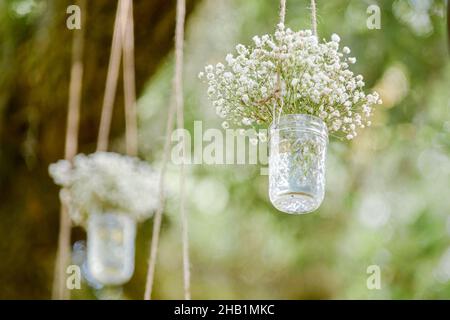 Mit Babys gefüllte Maurer-Gläser atmen bei der Hochzeitszeremonie im Freien Blumen, die an Bäumen hängen, gegen grünes Laub Stockfoto