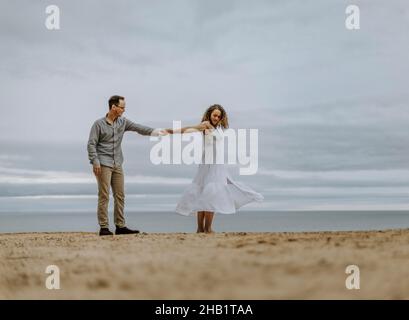 Mann tanzt mit Frau in weißem Kleid am Sandstrand neben dem Wasser Stockfoto