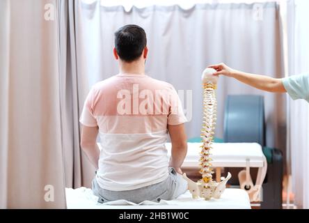 Mann, der auf dem Krankenhausbett saß und den Rücken drehte. Neben ihm Arzt hält Modell der Wirbelsäule. Stockfoto