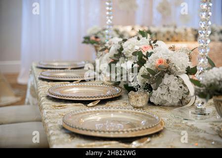 Wunderschöne Tischlandschaft gefüllt mit aufgereihten goldenen Ladegeräten, Blumen und Kristallvasen bei der Hochzeitsfeier Stockfoto