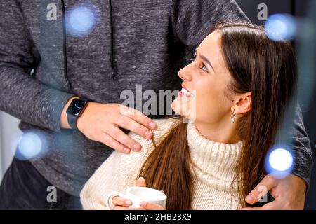 Fröhlich lächelnde junge Dame in weißem, warmen Pullover, die eine Tasse Tee in den Händen hält und ihren Freund an den Schultern umarmt. Stockfoto