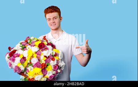 Glücklicher junger Mann, der einen riesigen schönen Blumenstrauß hält und mit seinem Zeigefinger auf blauem Hintergrund darauf zeigt. Stockfoto