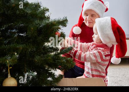 Kleines Mädchen hängt Dekorationen auf dem Weihnachtsbaum. Großmutter bewundert ihre Enkelin. Das Konzept der Familie und Neujahr Weihnachtsferien Stockfoto