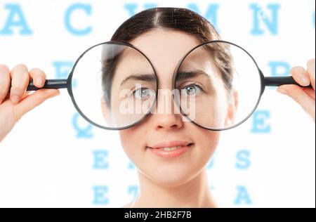 Augenheilkunde. Porträt einer jungen lächelnden hübschen Frau, die ihre Augen mit einer Lupe hält. Das Alphabet ist im Hintergrund. Das Konzept der Heilung Stockfoto
