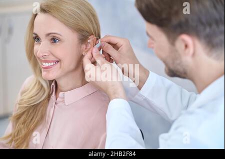 Montage. Mann in einem weißen Mantel, der Hörgeräte auf das Ohr einer lächelnden Frau mittleren Alters mit langen blonden Haaren im medizinischen Büro legt Stockfoto