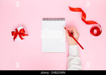 Weibliche Handschrift in leerem Notizbuch mit festlichen Verzierungen, Geschenkbox auf pastellrosa Hintergrund. Planung des neuen Jahres, Ziele, To-Do-Liste oder Wunschliste Stockfoto