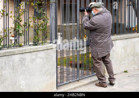 Ein Fotograf mit Corona Maske steht vor einem Metallzaun und fotografiert mit einem erweiterten Zoomobjektiv etwas auf dem Grundstück dahinter. Stockfoto