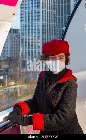 Junge Frau mit Gesichtsmaske während der Covid-19 Coronavirus-Pandemie, die im Dezember in London Eye Pod, Southbank, London UK, für Fotos posiert Stockfoto