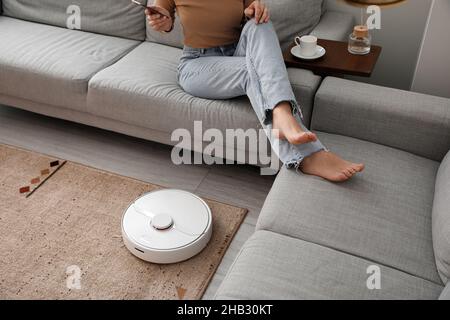 Moderner Roboter-Staubsauger in der Nähe des Sofas mit einer ruhenden Frau im Zimmer Stockfoto