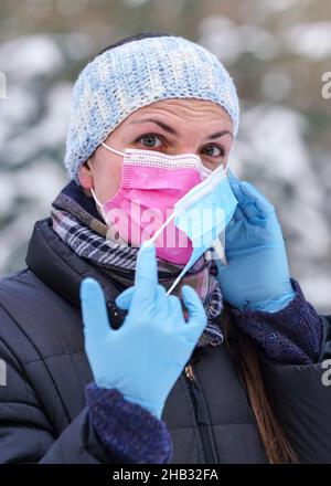 Junge Frau in warmer Winterkleidung trägt rosa Einweg-Virus Gesichtsmaske, setzen eine andere auf - einige raten, dass zwei Schichten bietet bessere Prot Stockfoto