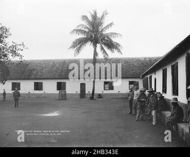 Gefängnis der Strafgefangenen, Inseln der Pines, Noumea, Studio der Burton Brothers, Fotostudio, 1899, Dunedin, Schwarz-Weiß-Fotografie, einstöckiges Kolonialgebäude mit Ziegeldach, das von vorne rechts nach hinten und dann über den Hintergrund verläuft. Ein offener Hof mit einem einfreien Kokosnussbaum in der Mitte im Hintergrund. Männer stehen und sitzen rechts an der Wand, ein Mann ist hinten in der Mitte und zwei andere Männer sind links hinten in der Nähe Stockfoto