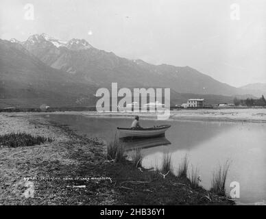 Glenorchy, Leiter des Lake Wakatipu, Burton Brothers Studio, Fotostudio, 1886, Dunedin, Schwarzweiß-Fotografie, Blick über einen ruhigen Teil des Wassers, auf dem ein Dingy mit einer einzigen Figur in der Mitte des Bildes sitzt. Es gibt schneebedeckte Berge in der Ferne auf der linken Seite und eine Siedlung jenseits des Wassers Stockfoto