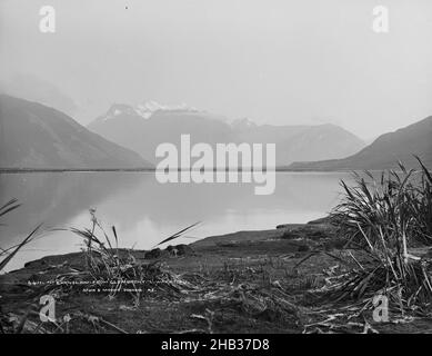 Mount Earnlsaw, von Glenorchy, Lake Wakatipu, Burton Brothers Studio, Fotostudio, 1886, Dunedin, Schwarz-Weiß-Fotografie, Blick über das stille Wasser eines Sees in Richtung schneebedeckter Berge. Auf dem Sandufer im Vordergrund sind zerklüllte Vegetationsklumpen zu sehen Stockfoto